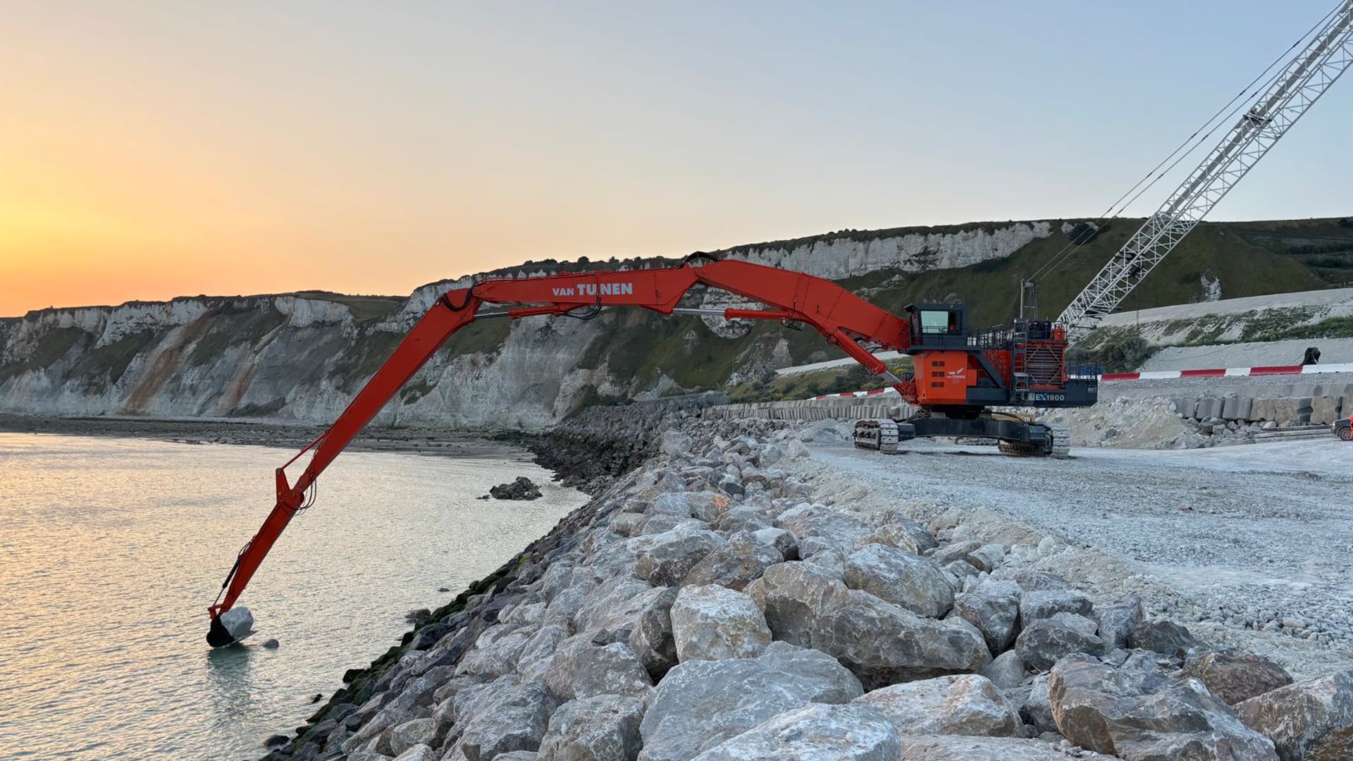 Long-reach excavator placing armour stone at the waterline, chalk cliffs behind
