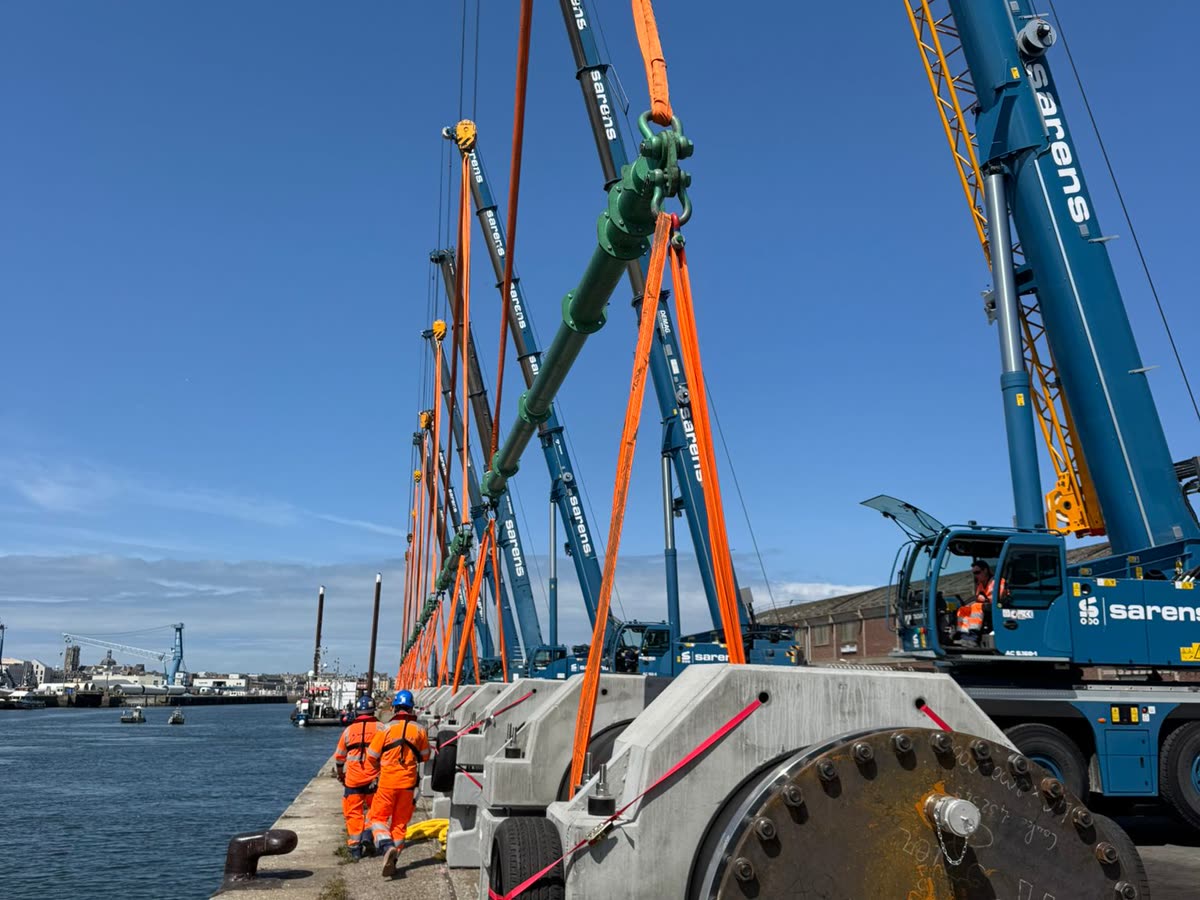 Several mobile cranes lifting a long flanged pipe on a quay